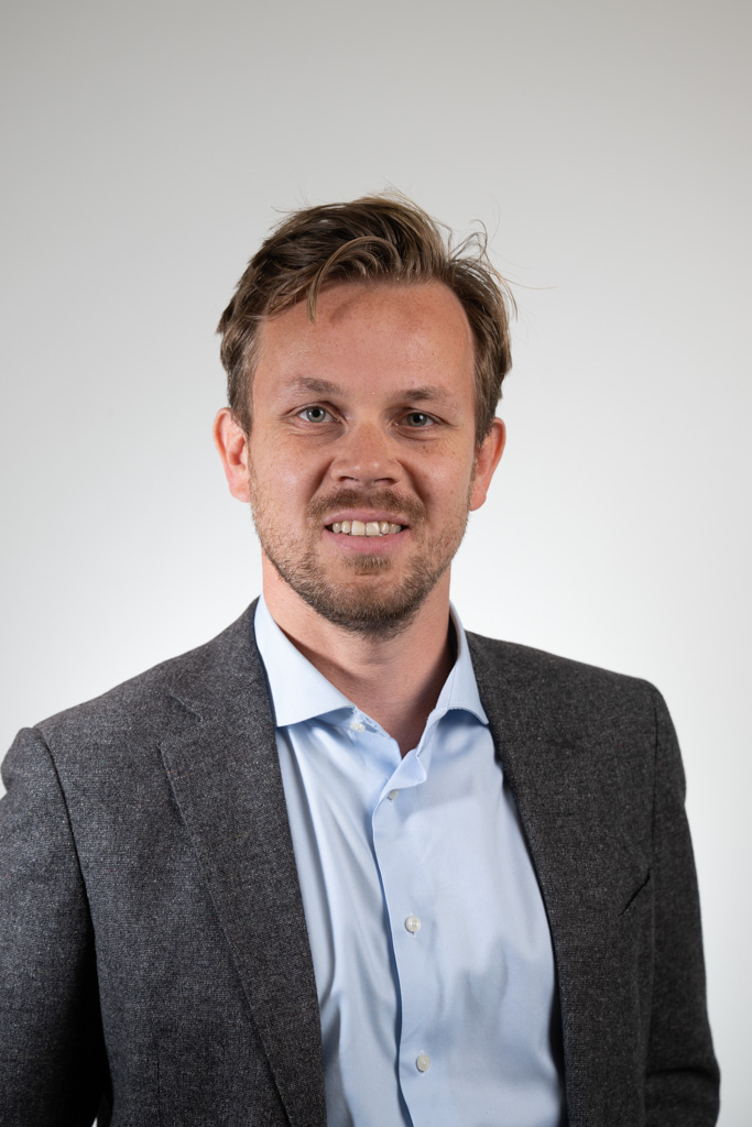 Portret van een glimlachende, zakelijk geklede man met kort bruin haar en baard, poserend tegen een neutrale witte achtergrond. De man draagt een lichtblauw overhemd onder een donkergrijs colbert, geschikt voor een professionele profielfoto of LinkedIn-portret.